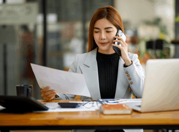 A woman sits at a desk reviewing a document while talking on the phone.