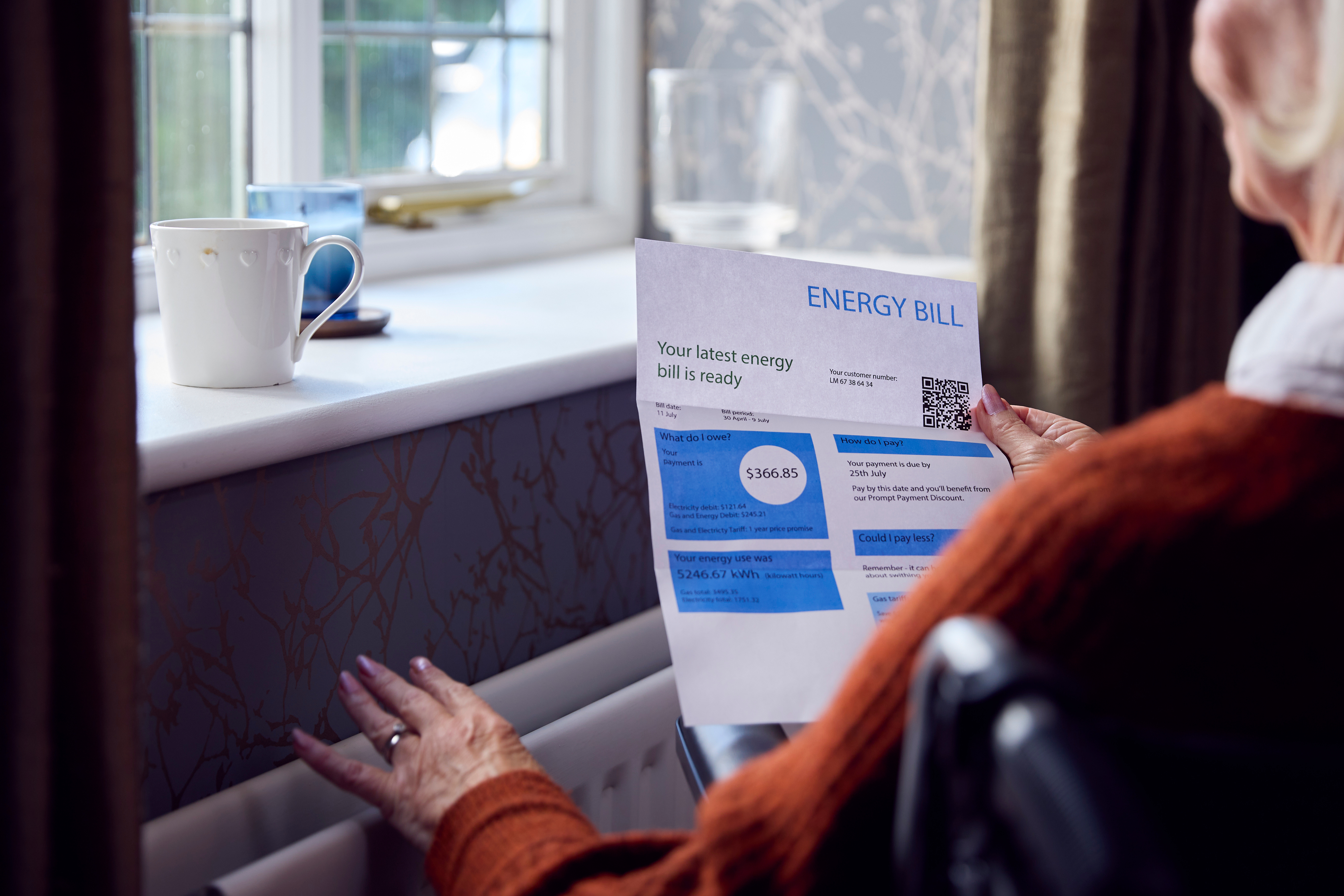 A person in a cozy room reads a printed energy bill while resting a hand on a radiator.