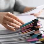 Hand organizing a large stack of documents secured with colorful binder clips on a desk.