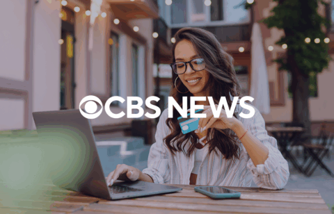 A person sitting outdoors uses a laptop while holding a credit card, with a CBS News logo overlaid on the image.