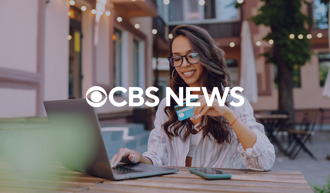 A person sitting outdoors uses a laptop while holding a credit card, with a CBS News logo overlaid on the image.