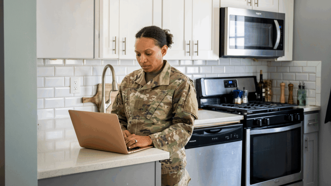 A woman in a military uniform stands at a kitchen counter using a laptop in her home.