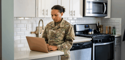 A woman in a military uniform stands at a kitchen counter using a laptop in her home.
