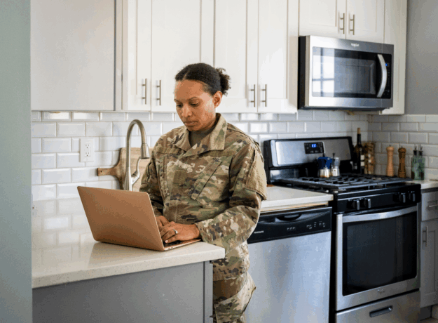 A woman in a military uniform stands at a kitchen counter using a laptop in her home.