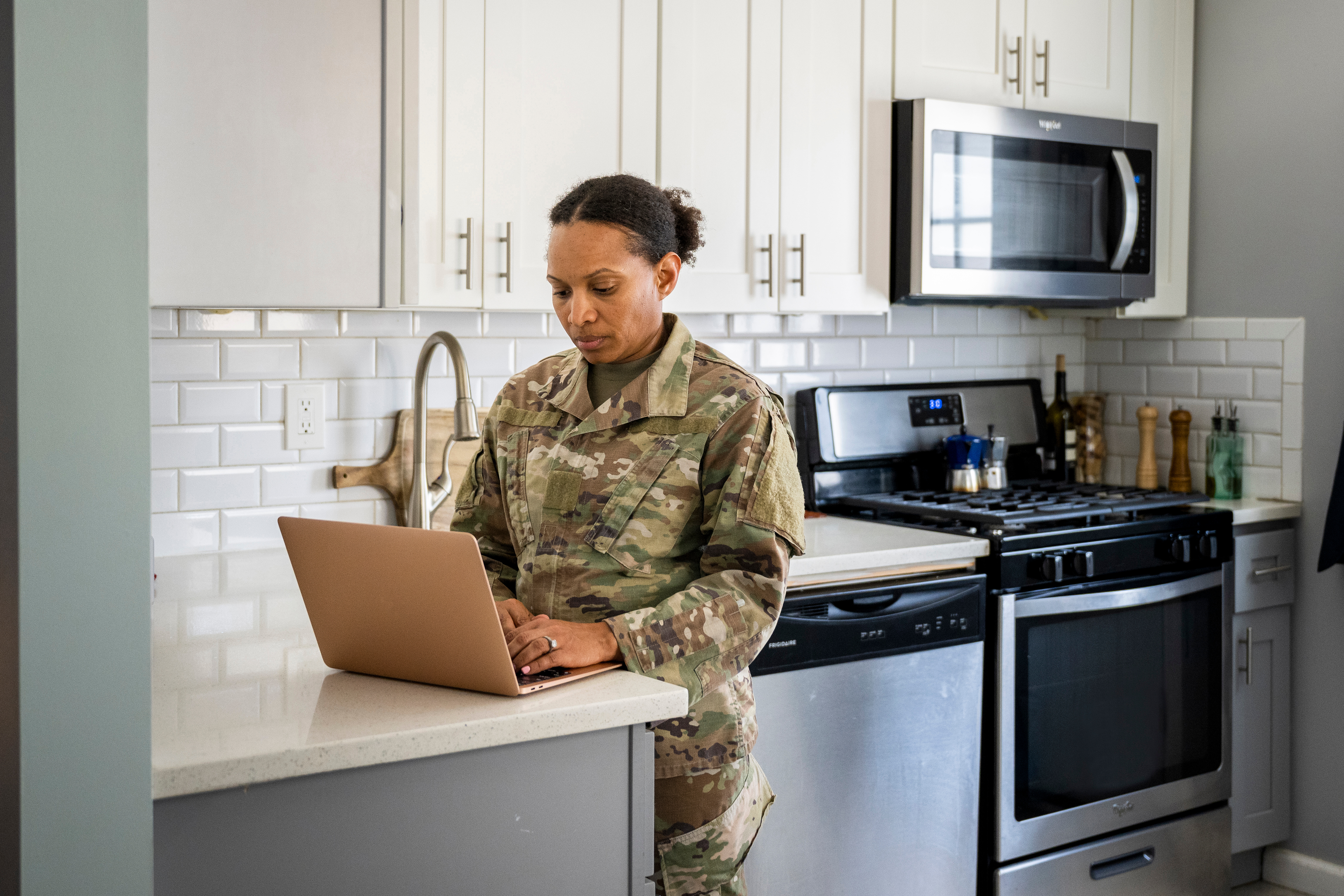 A woman in a military uniform stands at a kitchen counter using a laptop in her home.