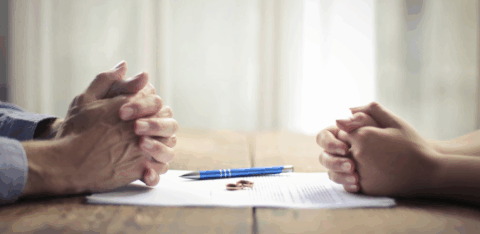 Two people sit across from each other at a table with clasped hands, a document, a pen, and wedding rings placed between them.
