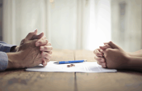 Two people sit across from each other at a table with clasped hands, a document, a pen, and wedding rings placed between them.