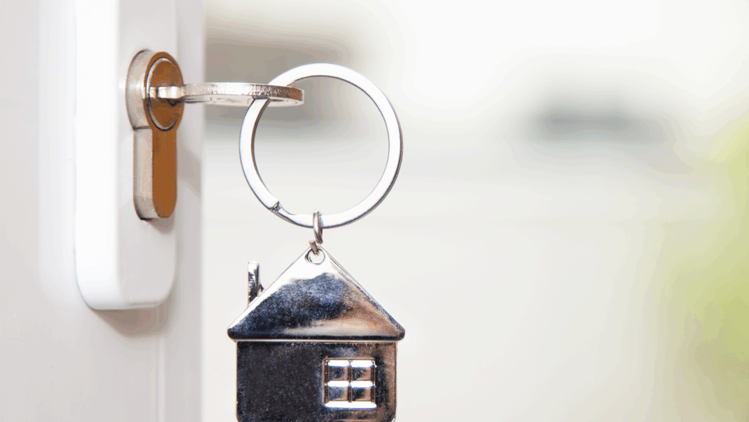 Close-up of a door key with a small metal house keychain attached, shown against a soft background.