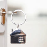 Close-up of a door key with a small metal house keychain attached, shown against a soft background.