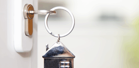 Close-up of a door key with a small metal house keychain attached, shown against a soft background.