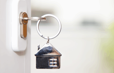 Close-up of a door key with a small metal house keychain attached, shown against a soft background.
