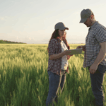 Two farmers stand in a green field reviewing information on a tablet during daylight.