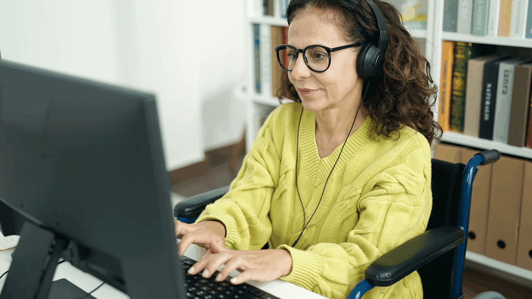 Woman wearing glasses and headphones uses a desktop computer while seated in a wheelchair in a home office.