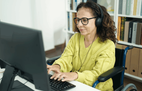 Woman wearing glasses and headphones uses a desktop computer while seated in a wheelchair in a home office.