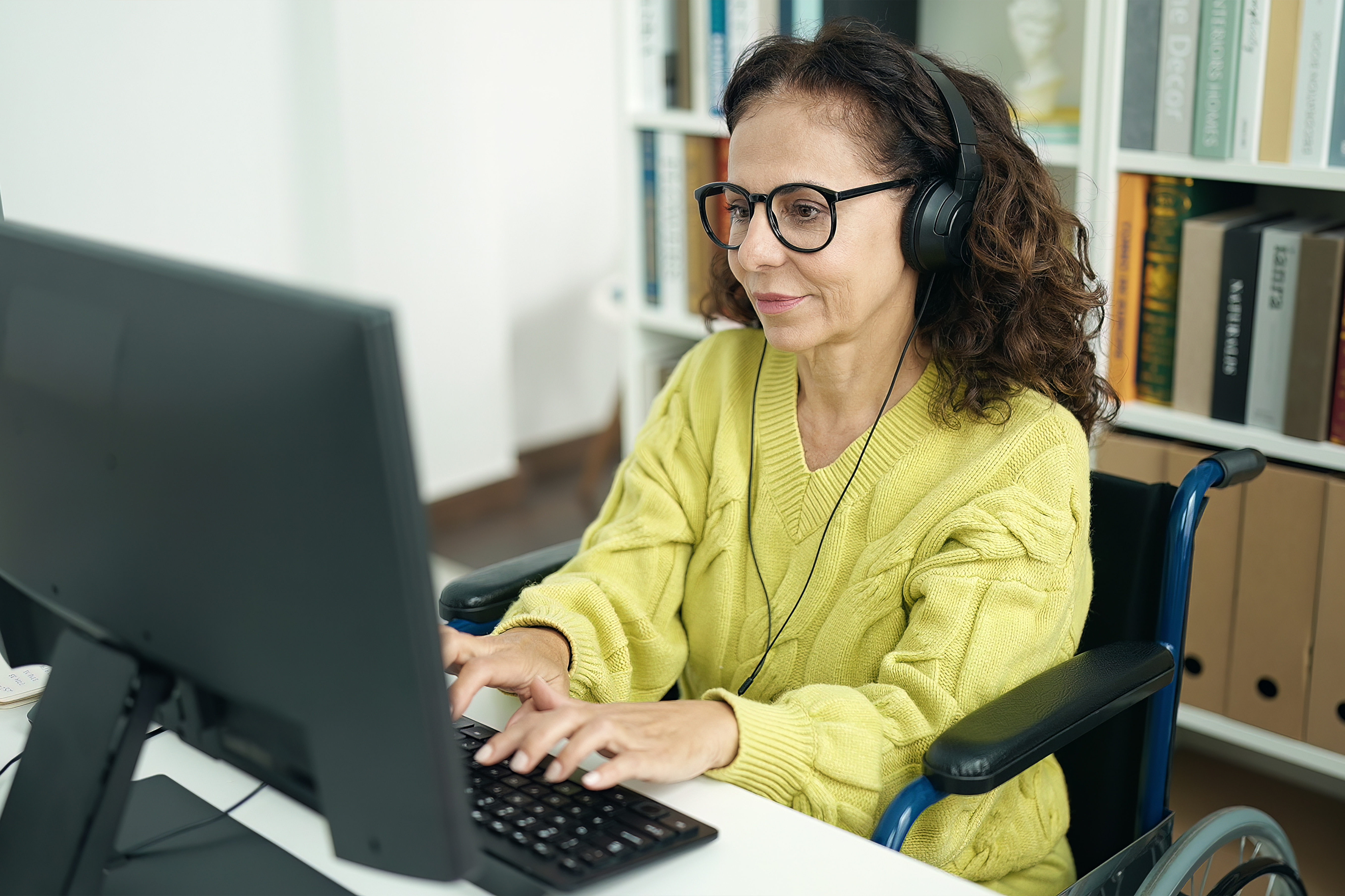 Woman wearing glasses and headphones uses a desktop computer while seated in a wheelchair in a home office.