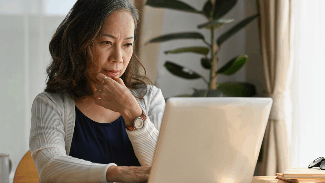 Older woman sitting at a table using a laptop, appearing focused while reviewing information at home.