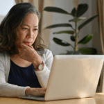 Older woman sitting at a table using a laptop, appearing focused while reviewing information at home.
