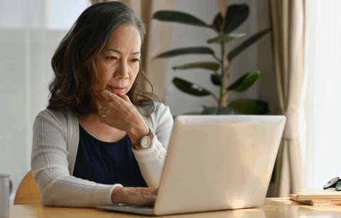Older woman sitting at a table using a laptop, appearing focused while reviewing information at home.