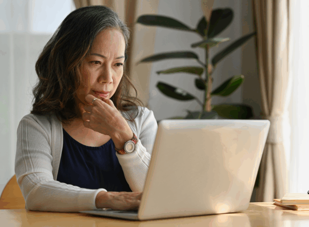 Older woman sitting at a table using a laptop, appearing focused while reviewing information at home.