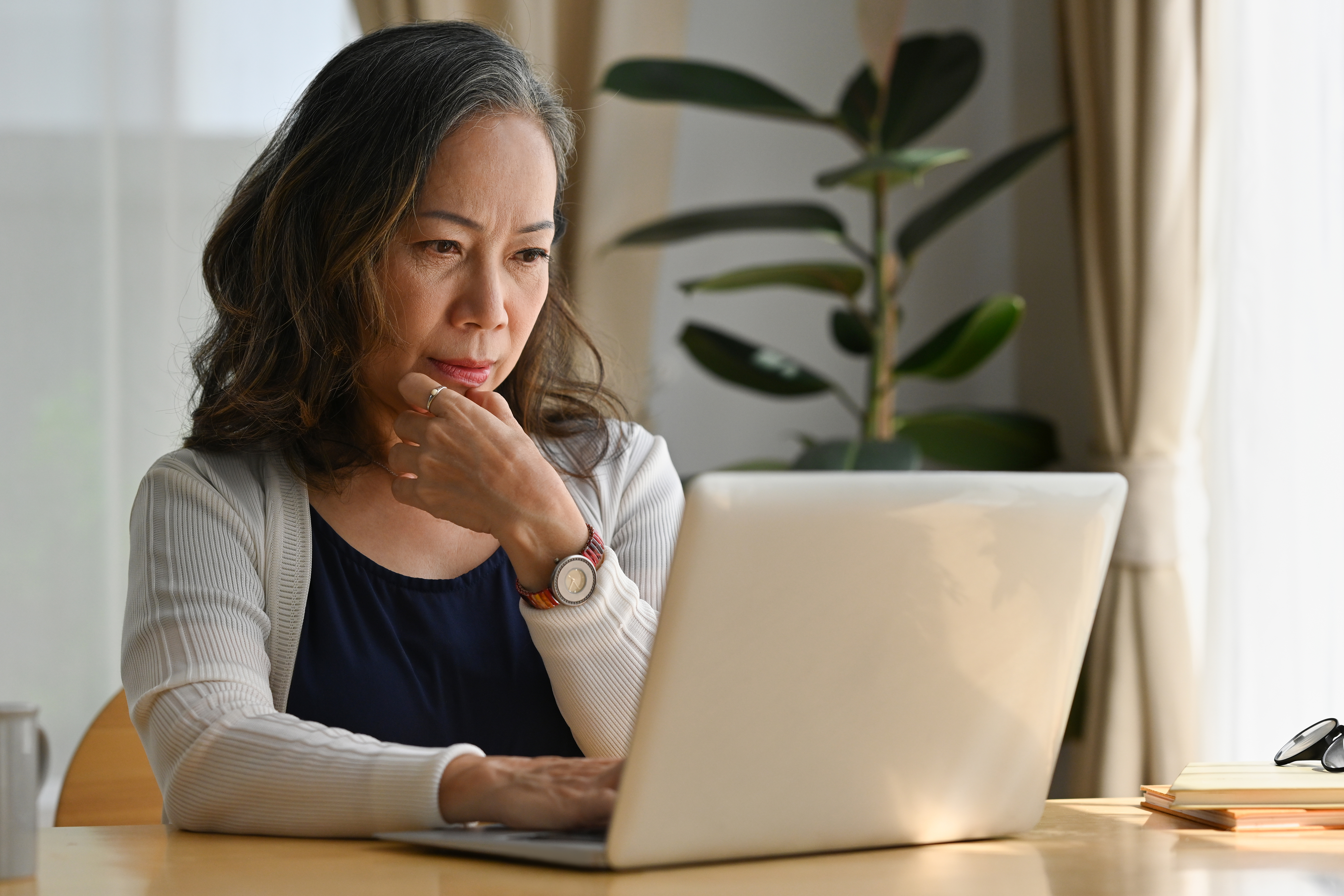 Older woman sitting at a table using a laptop, appearing focused while reviewing information at home.