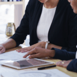 Two people reviewing paperwork together at a desk, with documents and a tablet laid out in front of them.