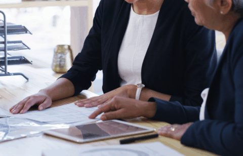 Two people reviewing paperwork together at a desk, with documents and a tablet laid out in front of them.