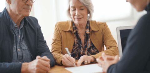 An older couple sits at a table with a professional, as one person signs a document while the others look on.
