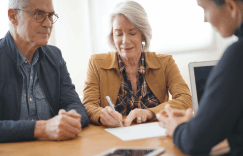 An older couple sits at a table with a professional, as one person signs a document while the others look on.