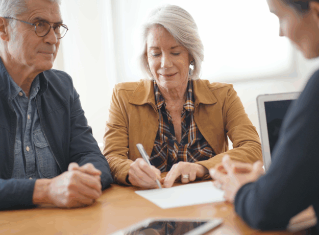 An older couple sits at a table with a professional, as one person signs a document while the others look on.