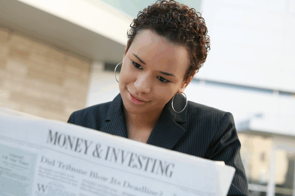 Woman in a business jacket reads a newspaper section titled “Money & Investing” while seated outdoors.