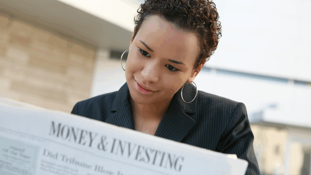Woman in a business jacket reads a newspaper section titled “Money & Investing” while seated outdoors.