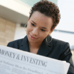 Woman in a business jacket reads a newspaper section titled “Money & Investing” while seated outdoors.