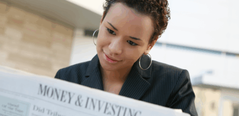 Woman in a business jacket reads a newspaper section titled “Money & Investing” while seated outdoors.