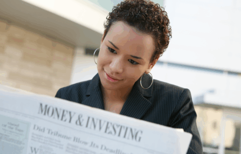 Woman in a business jacket reads a newspaper section titled “Money & Investing” while seated outdoors.