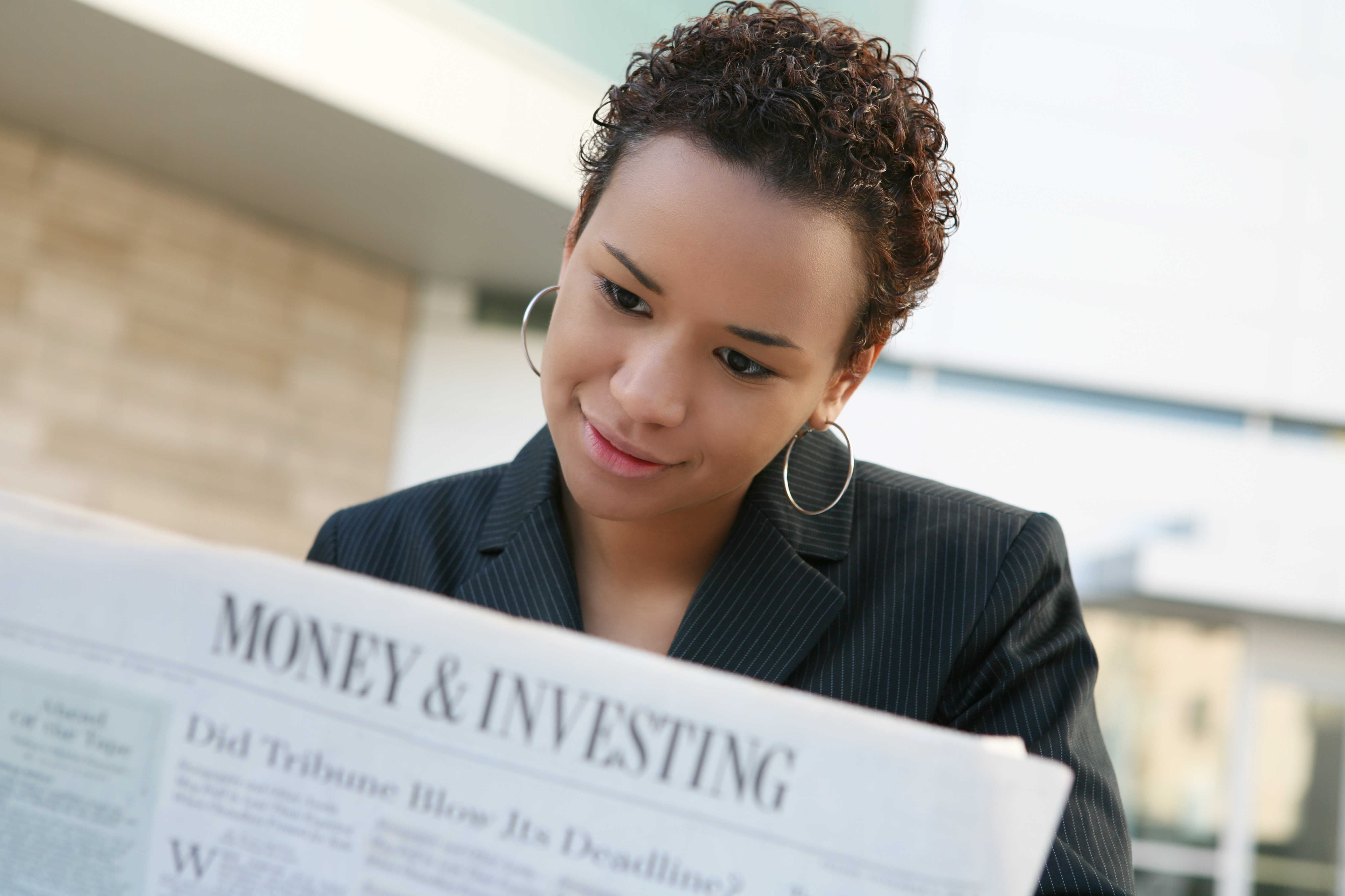 Woman in a business jacket reads a newspaper section titled “Money & Investing” while seated outdoors.