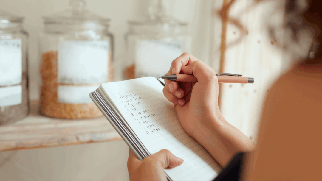 Person writing a grocery shopping list in a notebook with pantry jars in the background.