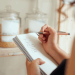 Person writing a grocery shopping list in a notebook with pantry jars in the background.