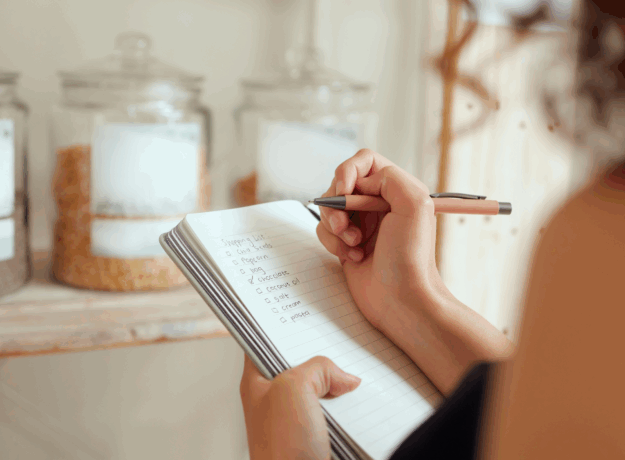 Person writing a grocery shopping list in a notebook with pantry jars in the background.