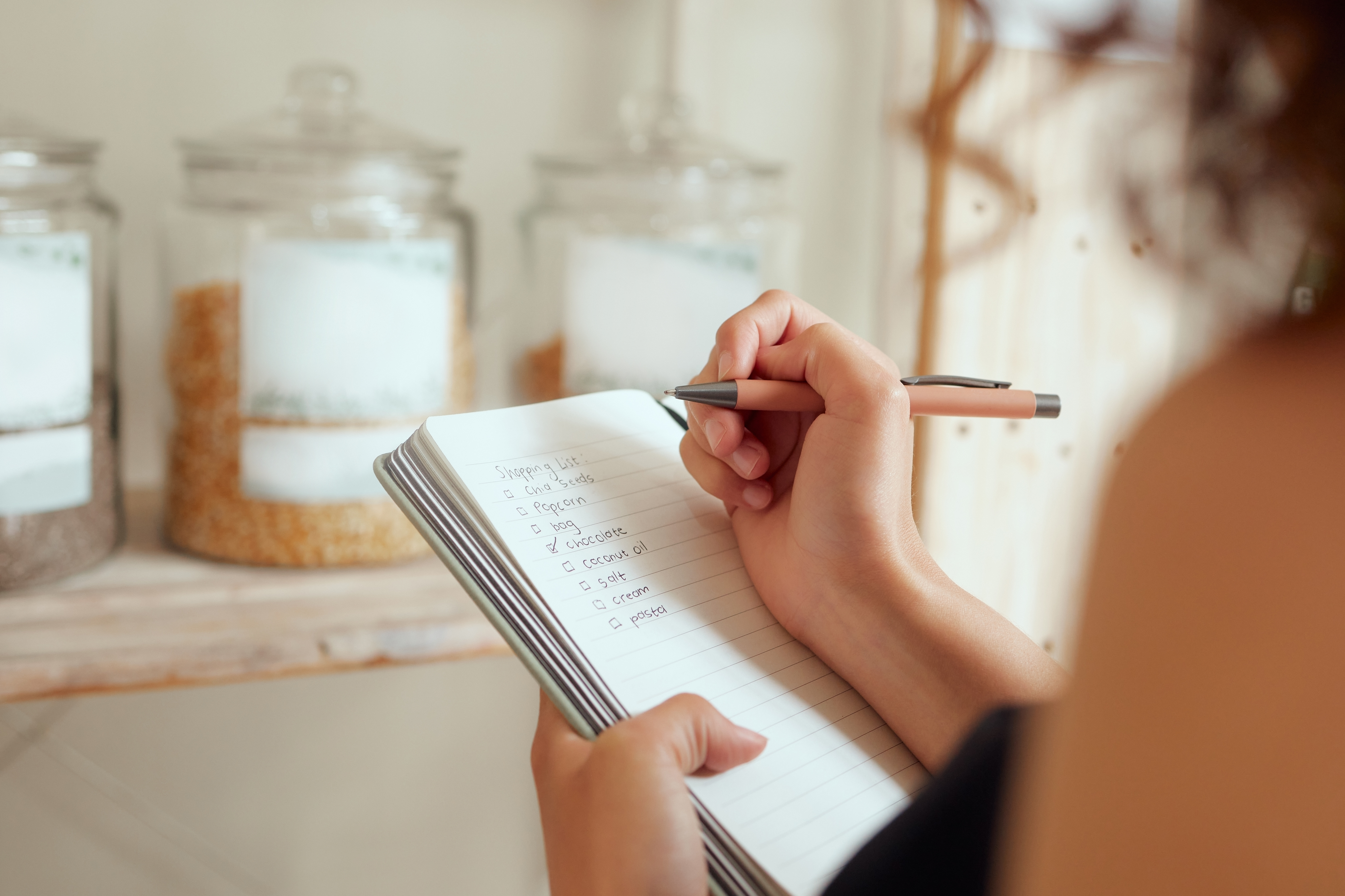 Person writing a grocery shopping list in a notebook with pantry jars in the background.
