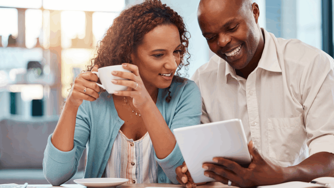 Couple sitting at a table reviewing financial documents on a tablet while drinking coffee at home.