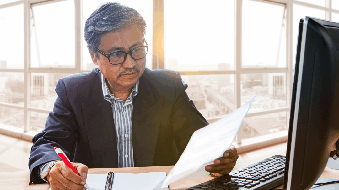 Professional reviewing documents while taking notes at a desk, with a desktop computer and papers nearby.