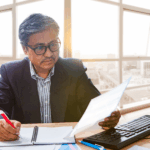 Professional reviewing documents while taking notes at a desk, with a desktop computer and papers nearby.