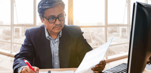 Professional reviewing documents while taking notes at a desk, with a desktop computer and papers nearby.