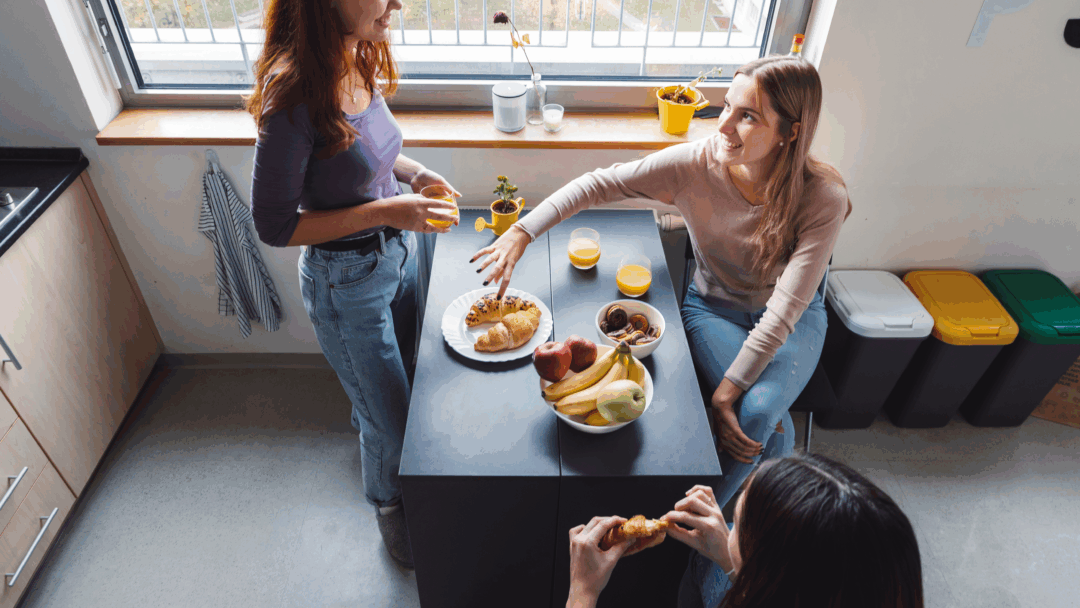 Three young adults sharing breakfast at a kitchen island with fruit, pastries, and drinks.