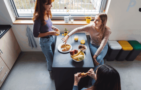 Three young adults sharing breakfast at a kitchen island with fruit, pastries, and drinks.