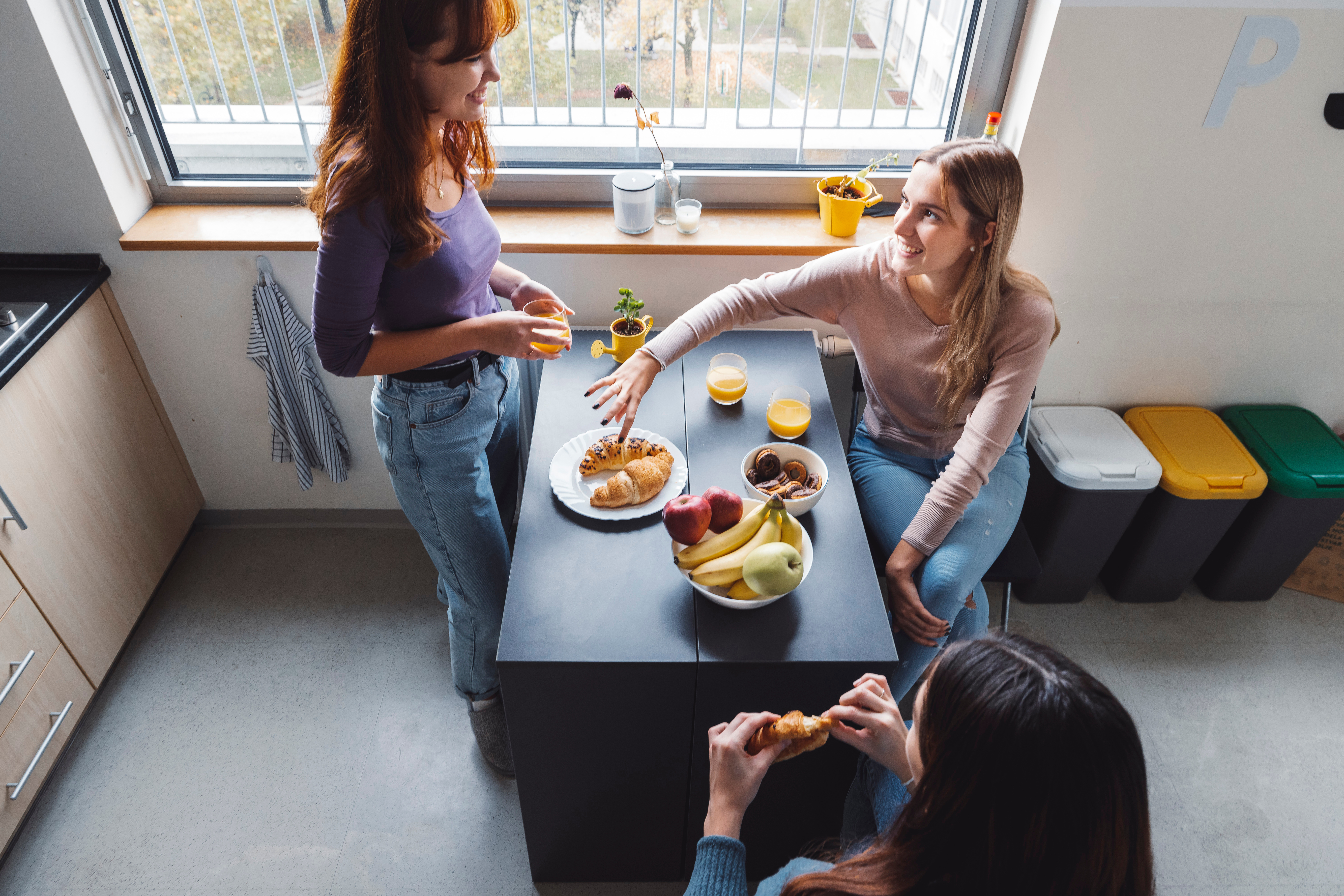 Three young adults sharing breakfast at a kitchen island with fruit, pastries, and drinks.