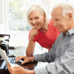 Older couple smiling while reviewing paperwork together at a home desk, with one person using a computer.