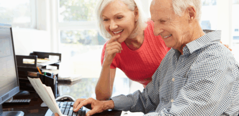 Older couple smiling while reviewing paperwork together at a home desk, with one person using a computer.