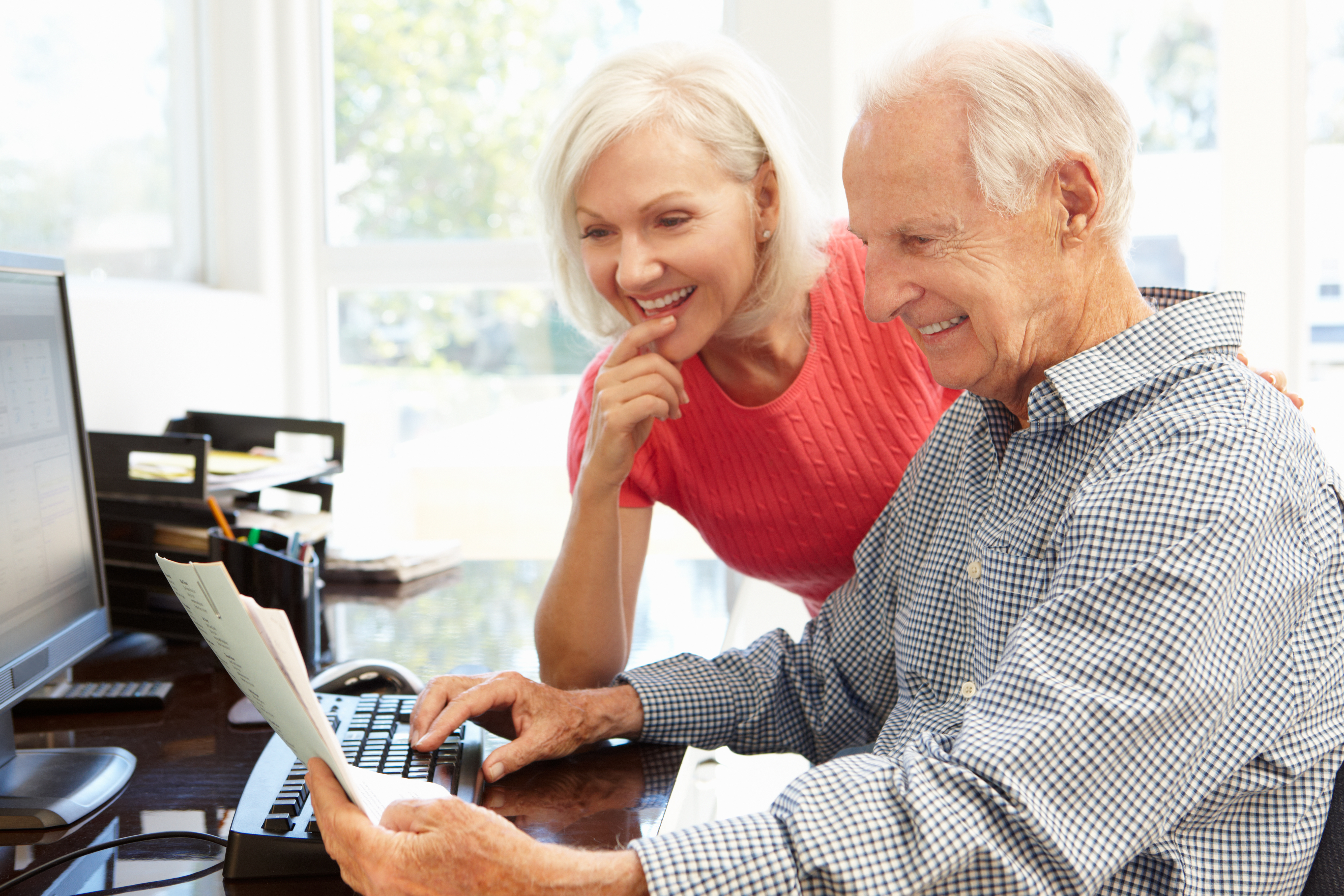 Older couple smiling while reviewing paperwork together at a home desk, with one person using a computer.