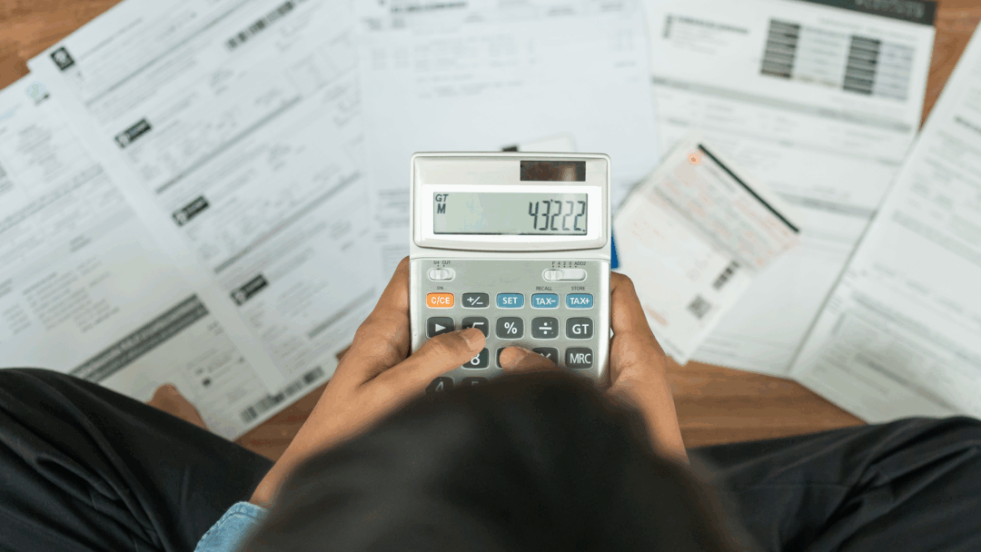 Person using a calculator while reviewing bills and financial documents spread across a desk.